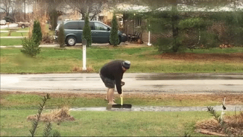 man with a hat stands next to a black car, holding the ground as he puts a blue marker into a hole in grass