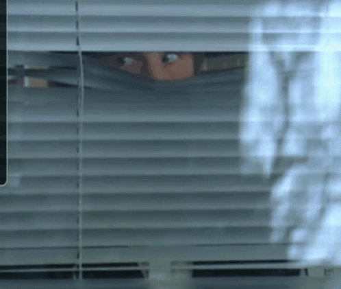 a woman looks out of the window with blinds
