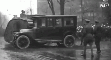 an old car traveling down a wet road with a lot of people standing in front of it