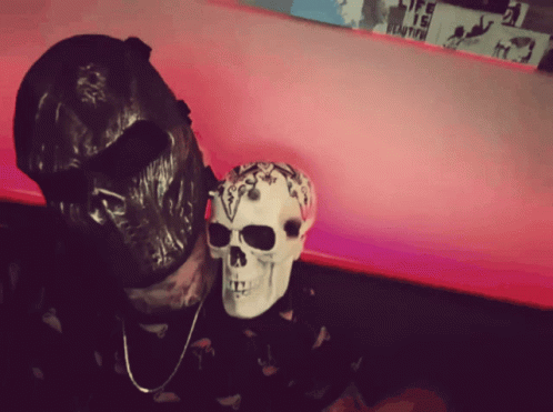 a man in skeleton mask and skull head covering sitting down next to a purple wall