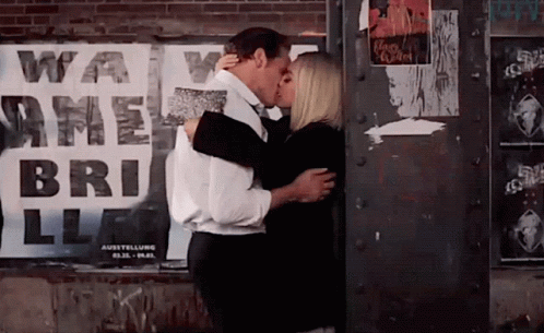 a couple kissing next to a wall covered with posters