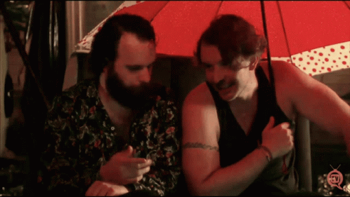 two men sitting under an umbrella looking at a cell phone