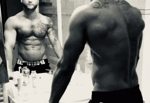 a man with tattoos standing in front of a mirror and looking at his reflection