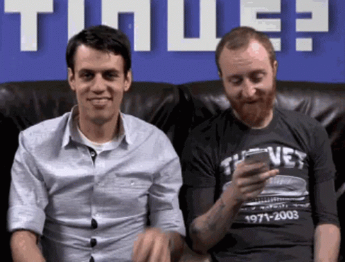 two men are sitting on a couch and one is texting on a cell phone