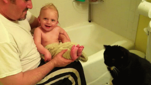 a man holding up a baby and cat next to a bath tub