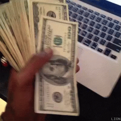 the person is holding a bunch of dollars in front of a laptop
