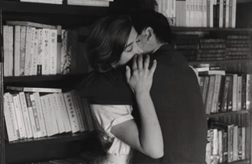 a man kisses his girlfriend as she stands in front of shelves full of books
