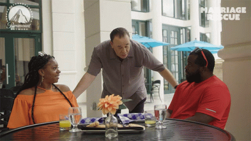 a man reaches out his arms to a table with two women