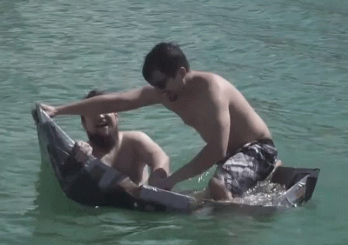 two shirtless guys play with a paper boat in the lake