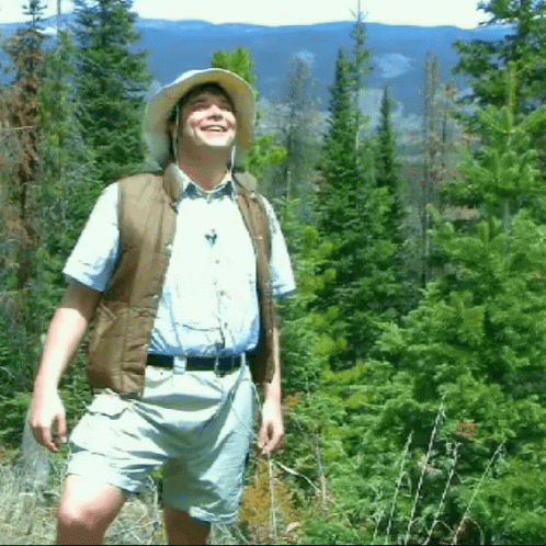 a man in a field of trees wearing a green hat and a gray vest