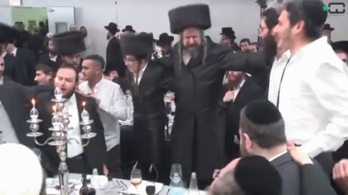 several people standing and sitting around with men in a jewish style room, one wearing a black hat