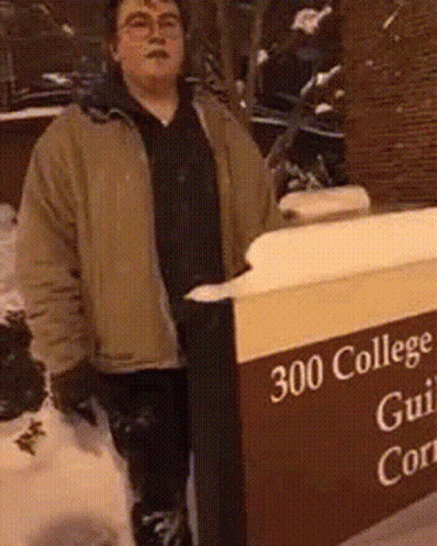 man wearing a gray jacket standing next to a sign for the college gullien corner