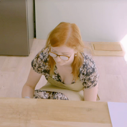 a woman in glasses and a blue dyed wig sits on the ground in front of a desk