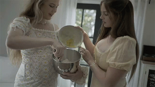 two women stand in a kitchen, one is holding a mixer and the other is looking at an object