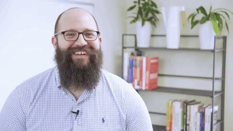 a man with beard wearing glasses stands in front of a bookshelf