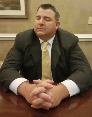 a man wearing a suit sitting at a table with his hands on his chest
