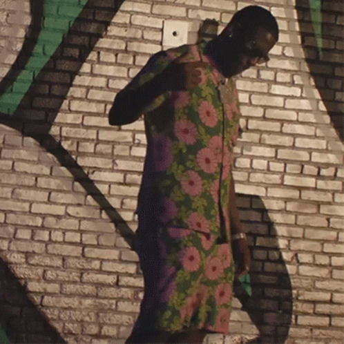 a man dressed in black and green and flowered clothing standing next to brick wall