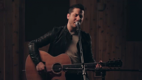 a man wearing a leather jacket sings into a microphone while holding an acoustic guitar