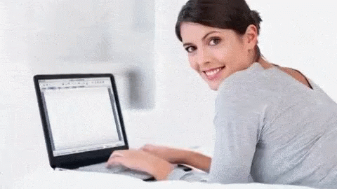 there is a smiling woman sitting on a bed using a laptop