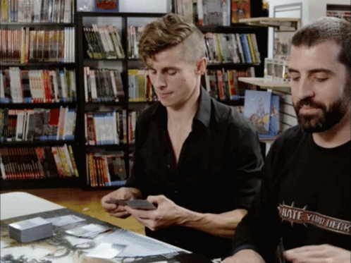 two men in front of bookshelves looking at electronics