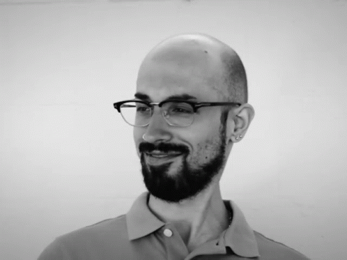 a bearded man in glasses and shirt looks at the camera