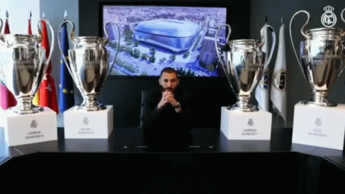 the man is sitting on a black table with five trophies behind him