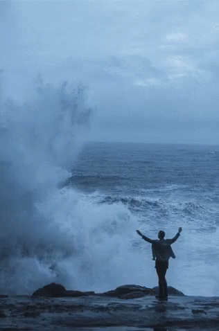 man standing on rock in front of large wave near shore