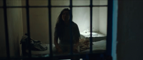 the girl stands in front of the bed behind bars in the cell