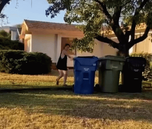 a woman walking across a yard to some trashcans