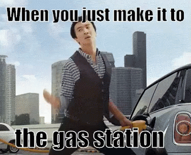 a man wearing an apron, holding up an air freshener, and jumping out of a car in a gas station