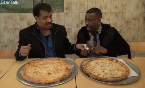 two men are sitting in front of two large pizzas