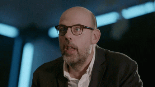 a bald man in glasses looks on as he talks