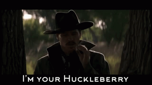a man wearing a hat standing in a forest with an i'm your huckleberry written across it