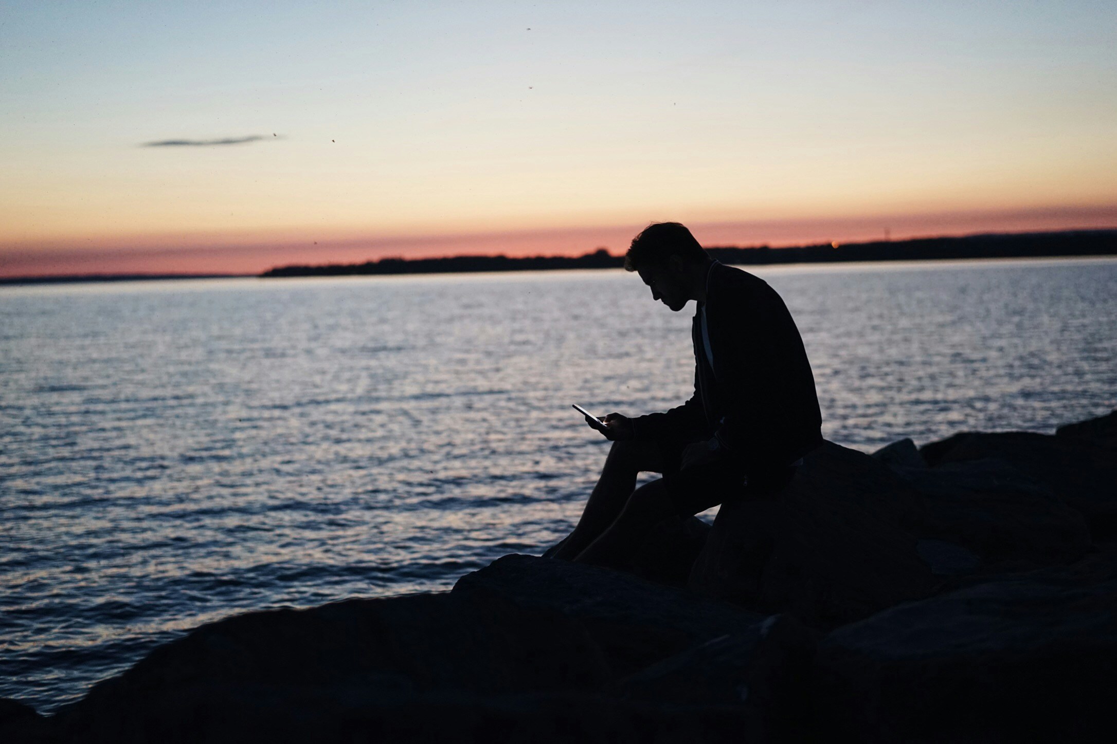 a man sitting on a rock on the ocean watching a screen